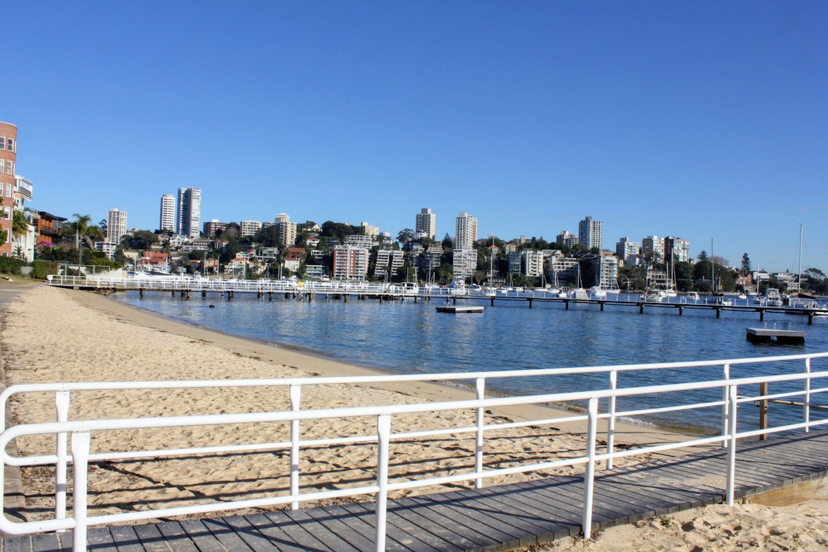 Sandy beach with white railings in the foreground, marina with boats, and city skyline with high-rise buildings under a clear blue sky.