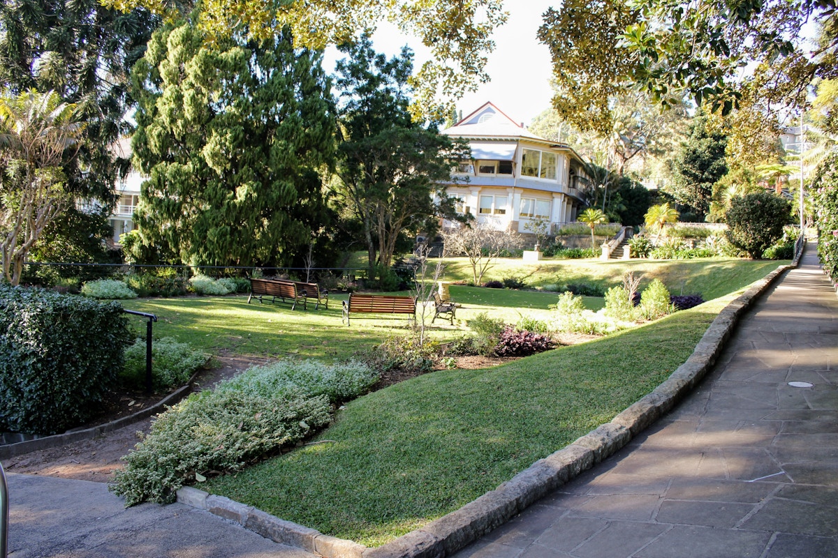 A paved pathway runs beside a landscaped garden with benches, shrubs, and trees, leading toward a large house in the background on a sunny day.