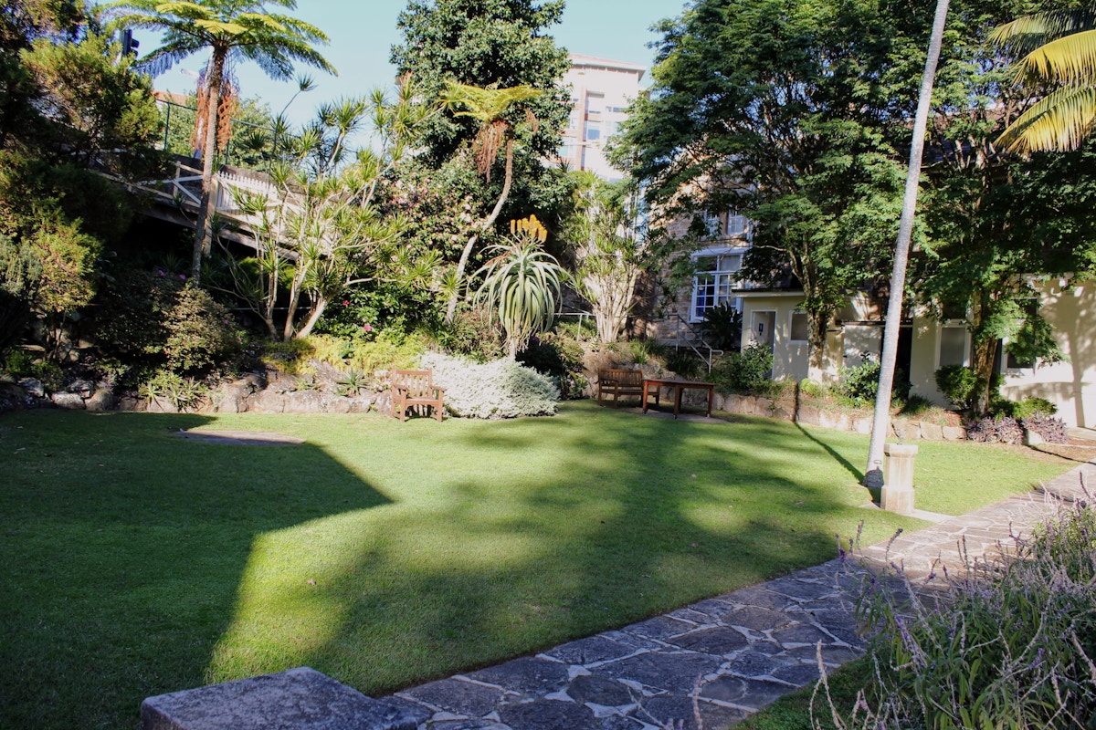 Small garden with lawn, benches, stone path, and various trees and shrubs in sunlight. Building and lamppost visible in the background.
