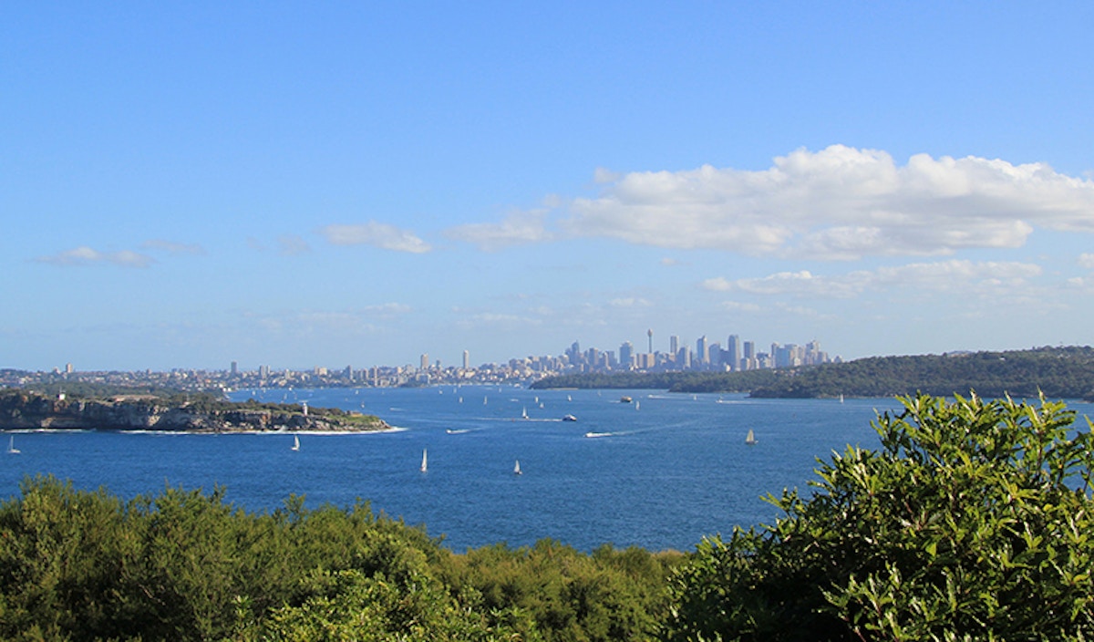 View of a blue harbor with sailboats, green islands in the foreground, and a city skyline with tall buildings under a clear, partly cloudy sky in the distance.
