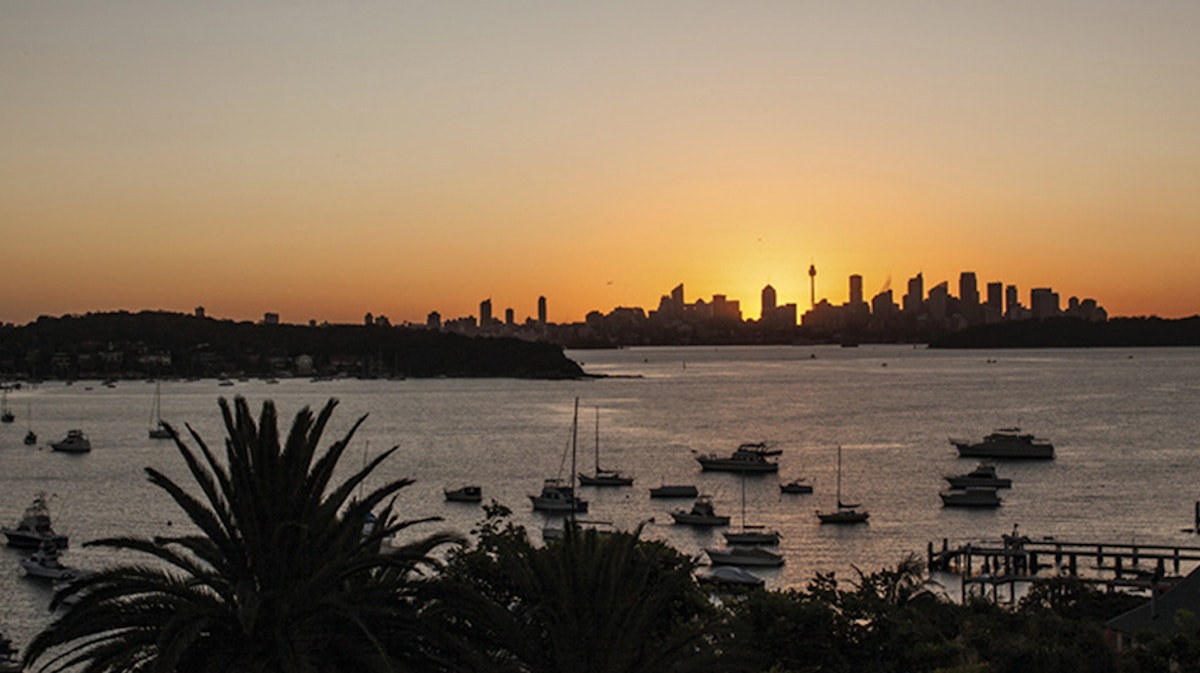 Boats anchored in a harbor at sunset, with city skyline and tall buildings silhouetted against an orange sky in the background.