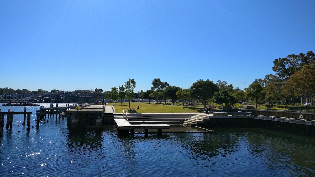 A waterfront park with grassy areas, trees, steps, and boat docks under a clear blue sky.