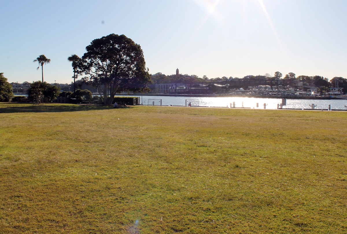 A grassy field near a river with trees, buildings, and boats in the background under a bright, sunny sky.