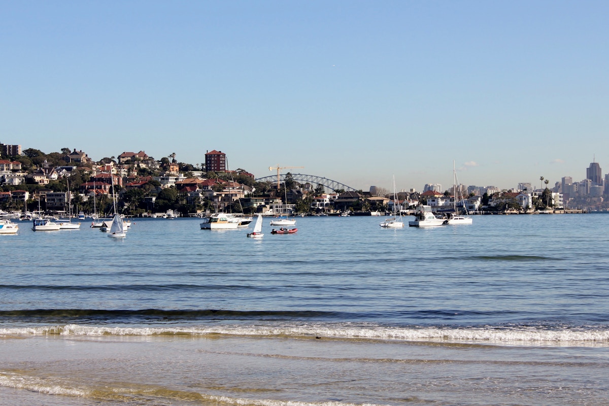 Boats are anchored in a bay with houses on a hill and a bridge in the background under a clear sky.