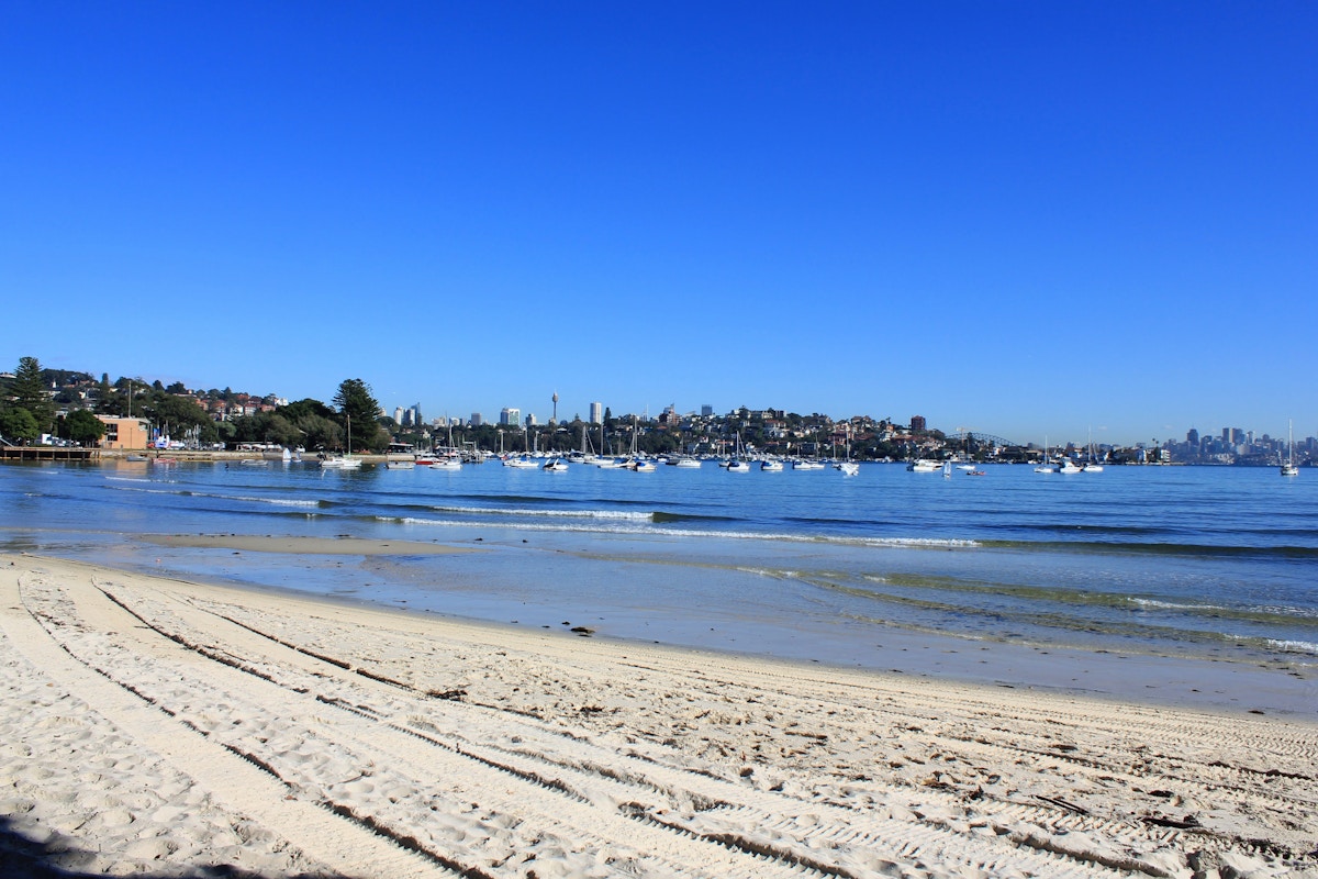 Sandy beach with tire tracks in the foreground, calm water with anchored boats, and a city skyline in the background under a clear blue sky.