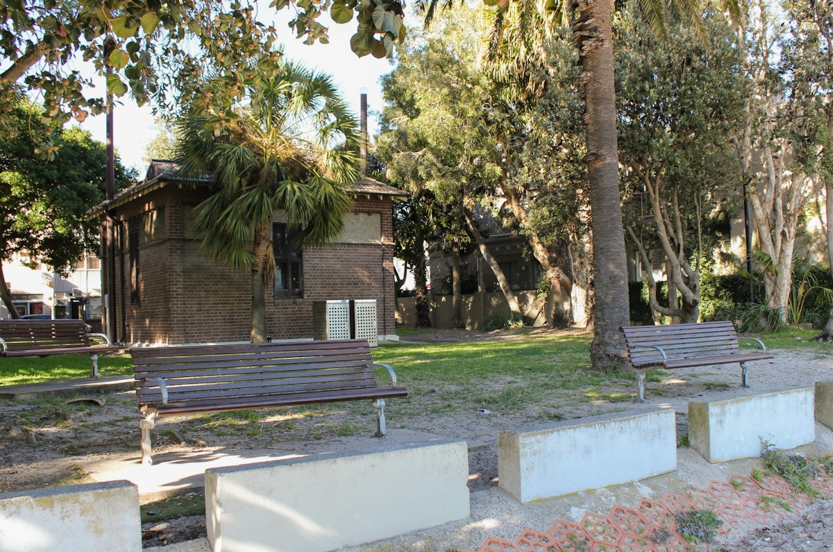 Two empty wooden benches face a brick building in a shaded park area with trees and concrete barriers.