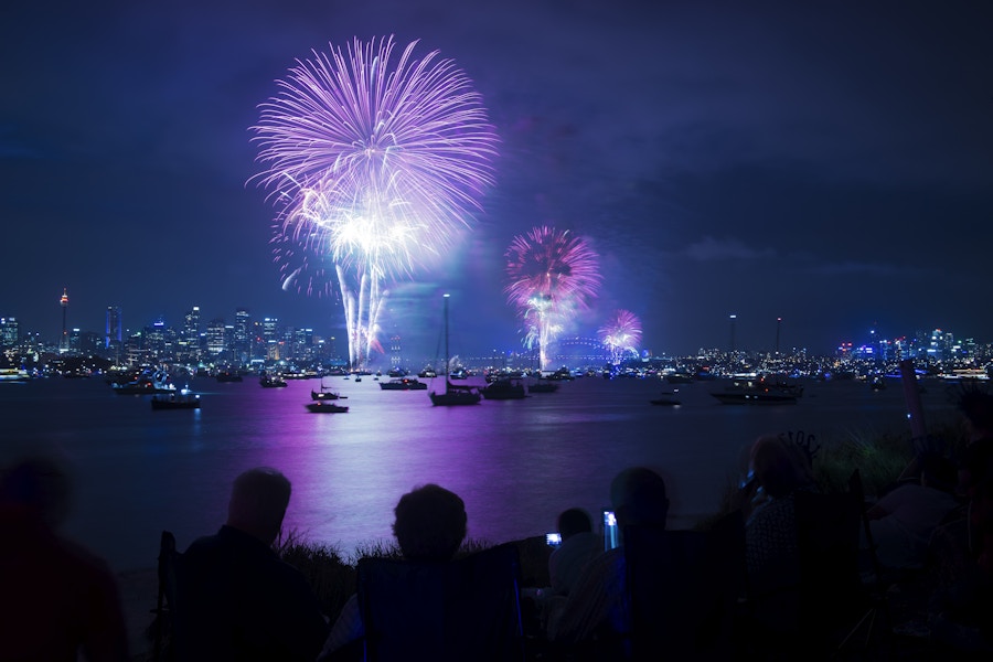 People watch colorful fireworks lighting up the night sky over a city skyline, with reflections on the water and boats in the harbor.