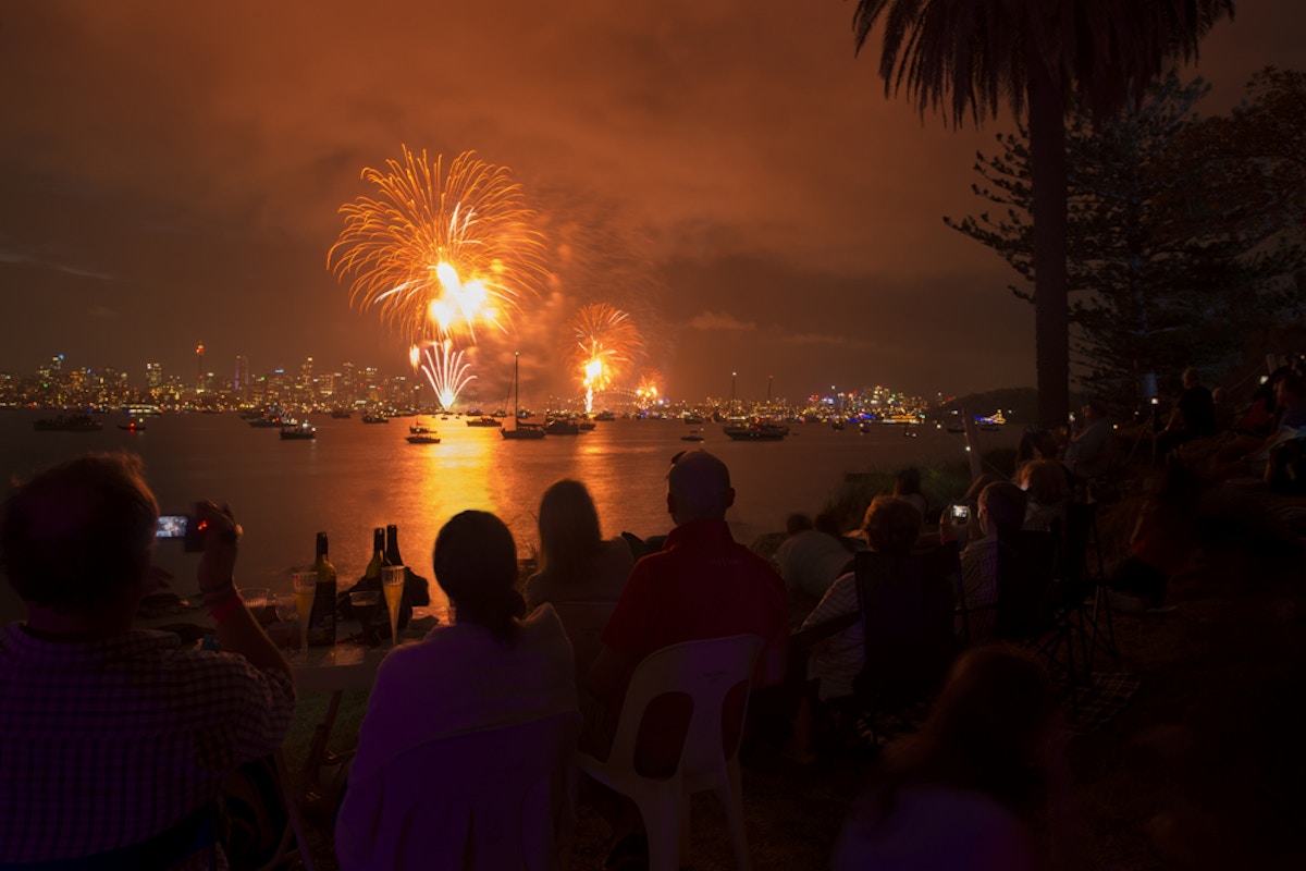 People sit by the water at night, watching bright fireworks explode over a city skyline. Boats are on the water and trees frame the scene.
