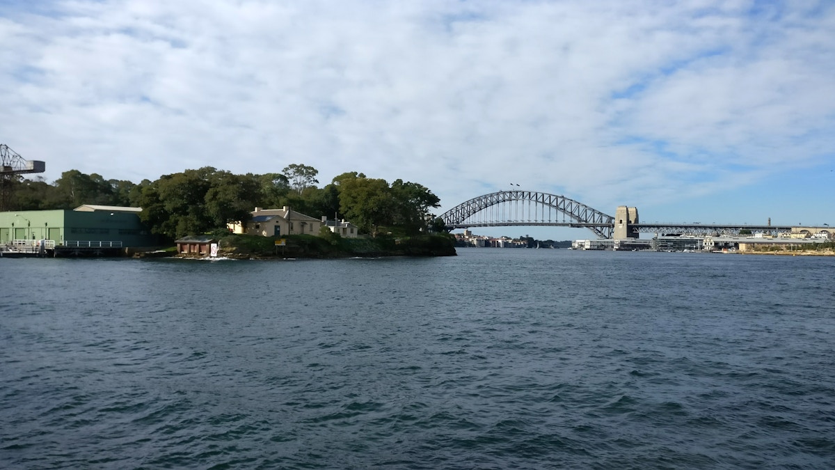Sydney Harbour with an island in the foreground, water in the center, and the Sydney Harbour Bridge in the background under a partly cloudy sky.