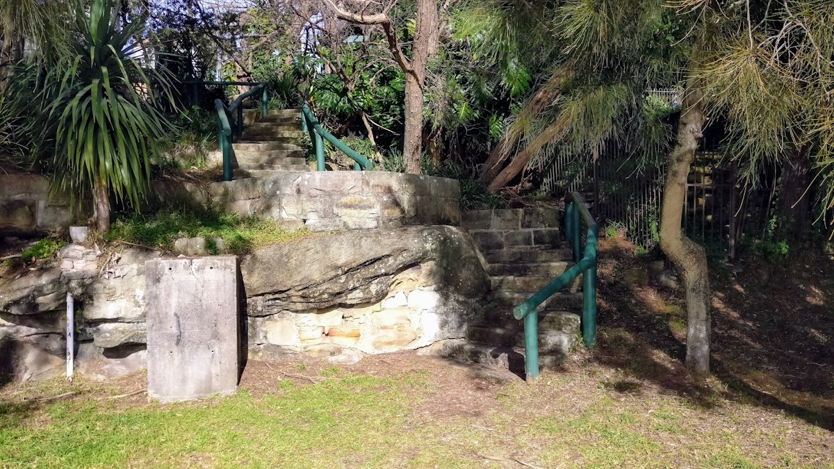 Stone steps with green railings ascend a small grassy hill, surrounded by rocks, trees, and dense greenery in bright sunlight.