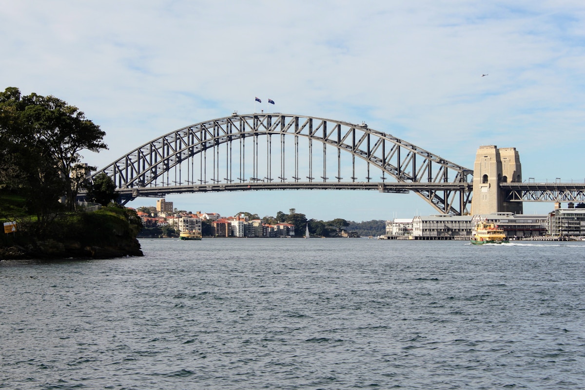 Sydney Harbour Bridge spans over the water with a ferry passing below; city buildings and greenery visible on both sides under a partly cloudy sky.