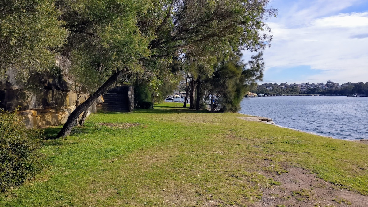 Grassy area with trees beside a calm body of water, a stone wall to the left, and houses visible across the water under a partly cloudy sky.