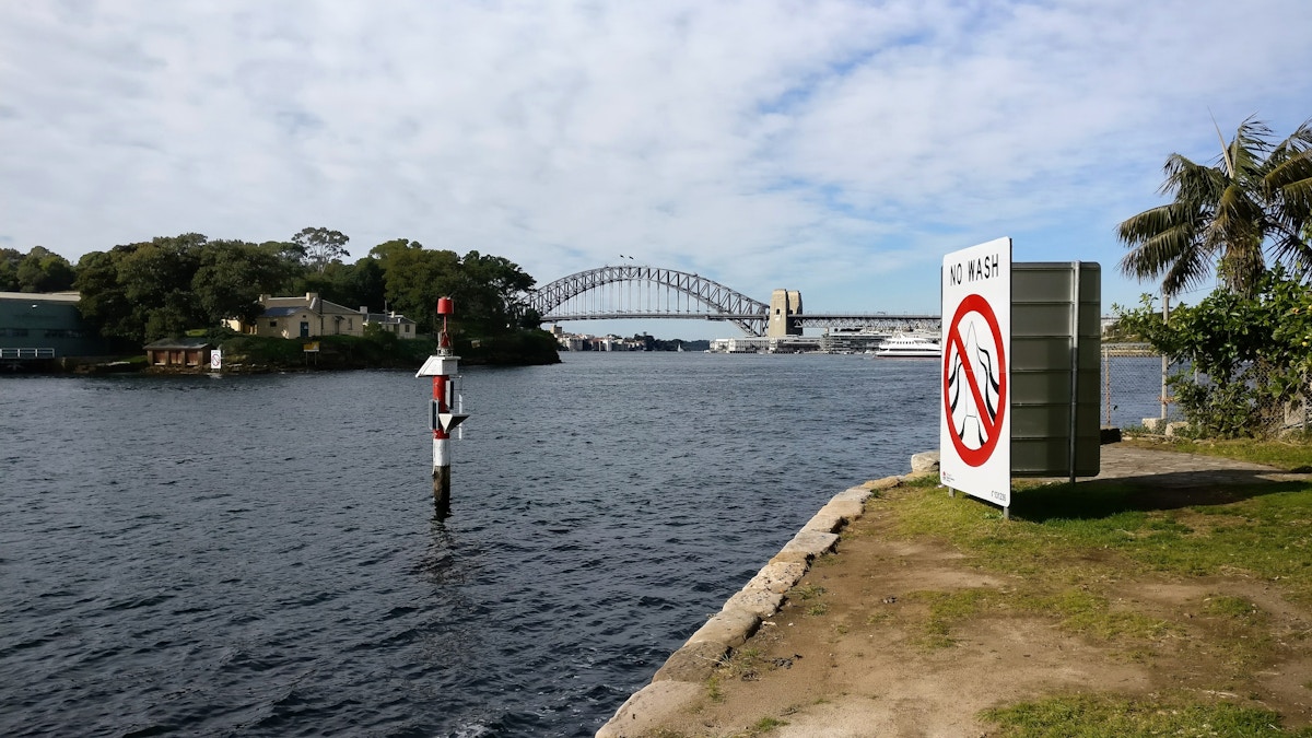 A body of water with a no fishing sign in the foreground, a navigational marker in the water, and a large bridge in the background under a partly cloudy sky.