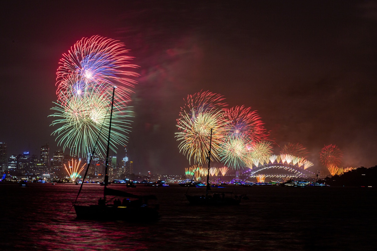 Colorful fireworks light up the night sky over Sydney Harbour, with boats visible on the water and the Sydney Opera House in the background.