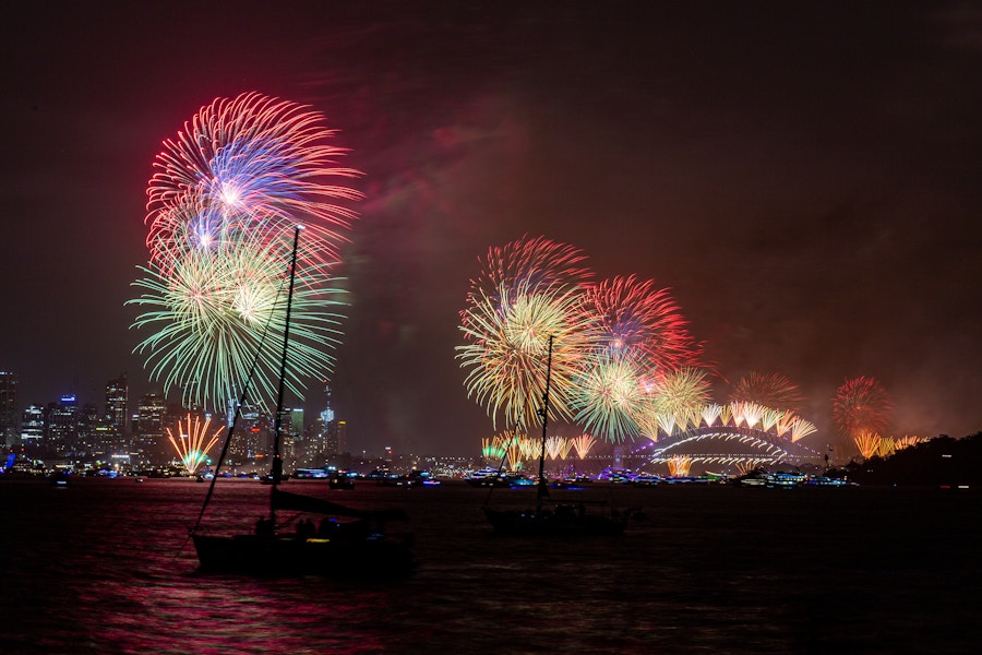 Colorful fireworks light up the night sky over Sydney Harbour, with boats visible on the water and the Sydney Opera House in the background.