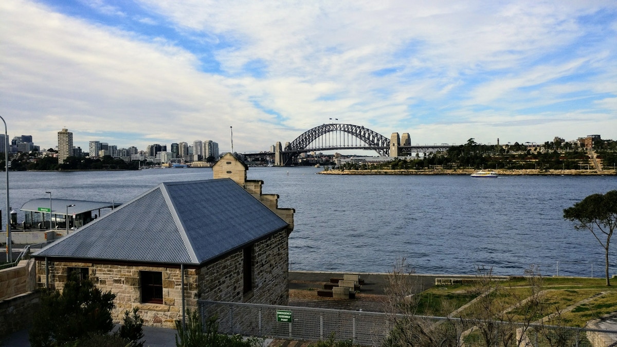 A cityscape featuring the Sydney Harbour Bridge over the harbor, with a stone building and greenery in the foreground under a partly cloudy sky.