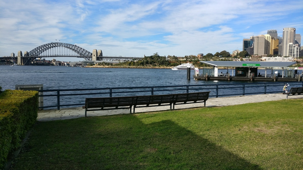 Benches overlook Sydney Harbour with the Sydney Harbour Bridge on the left, city buildings on the right, and a ferry wharf along the waterfront under a partly cloudy sky.