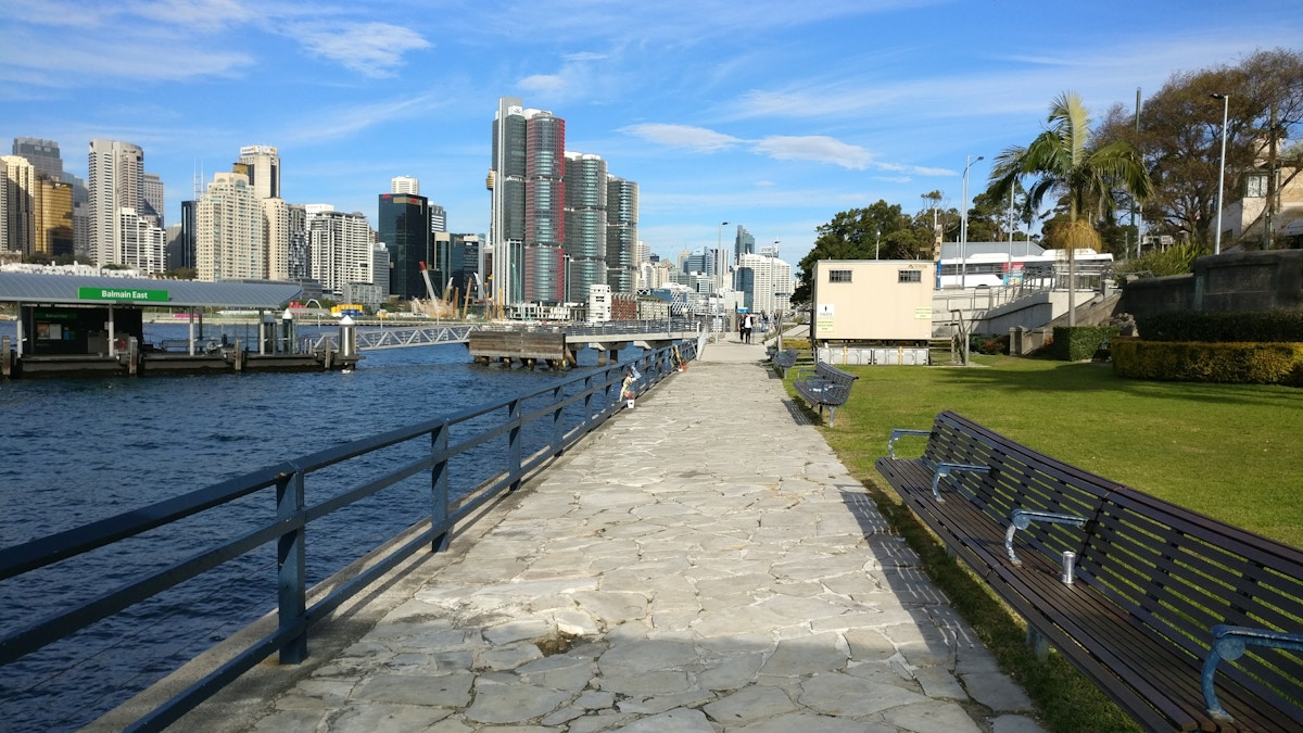 Stone walkway along a waterfront with city skyscrapers in the background, benches and green lawn on the right, and blue sky above.