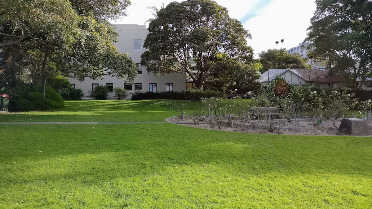 Green lawn with a small garden bed, trees, and two buildings in the background under a partly cloudy sky.