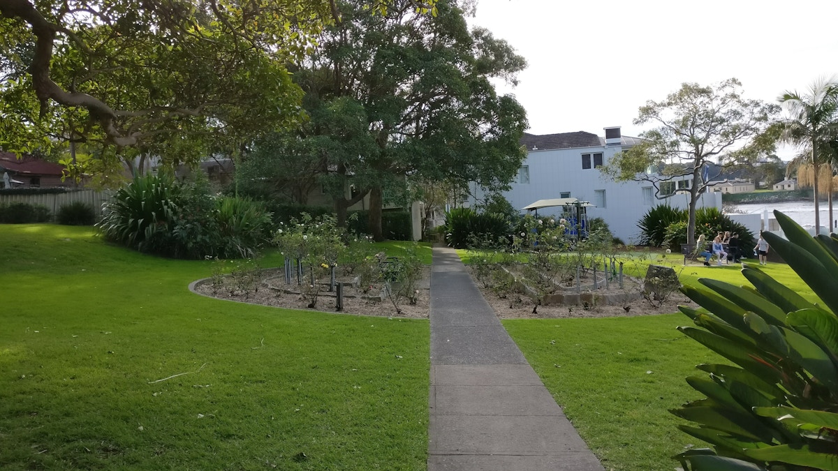A paved path leads to a circular garden bed surrounded by grass, with houses, trees, and people sitting near the water in the background.