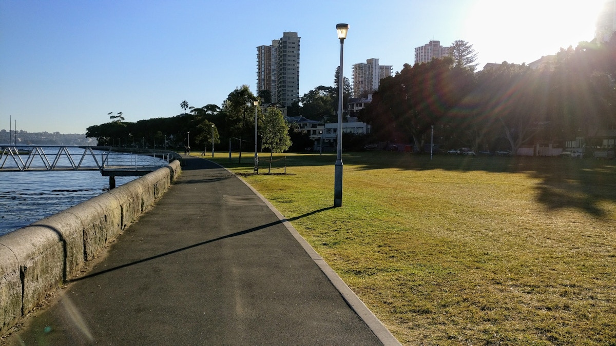 A paved path runs beside the water with grass, trees, and tall buildings in the background on a sunny day.