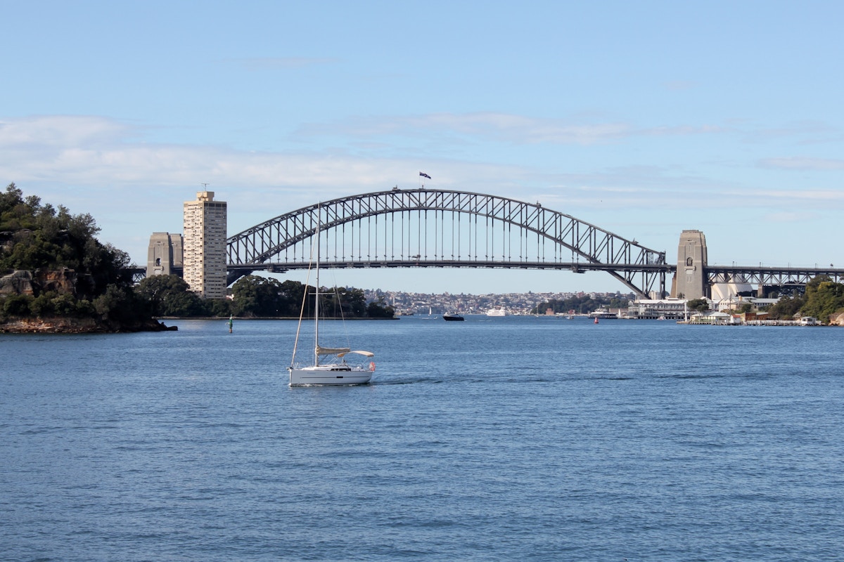 A sailboat is on the water with the Sydney Harbour Bridge and city skyline in the background under a clear sky.