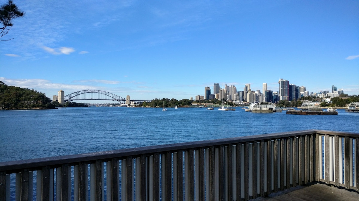 View of a city skyline and harbor with boats, including a bridge on the left, seen from behind a wooden railing on a clear day.