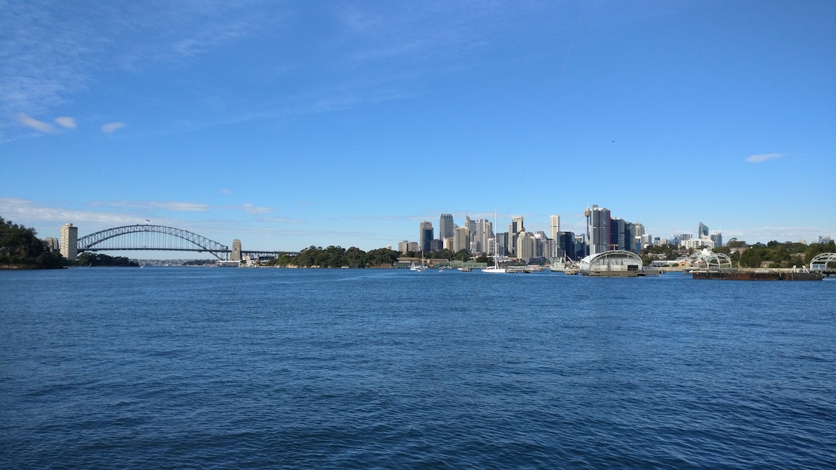 Sydney skyline with tall buildings, Sydney Harbour Bridge on the left, blue sky, and water in the foreground under bright daylight.