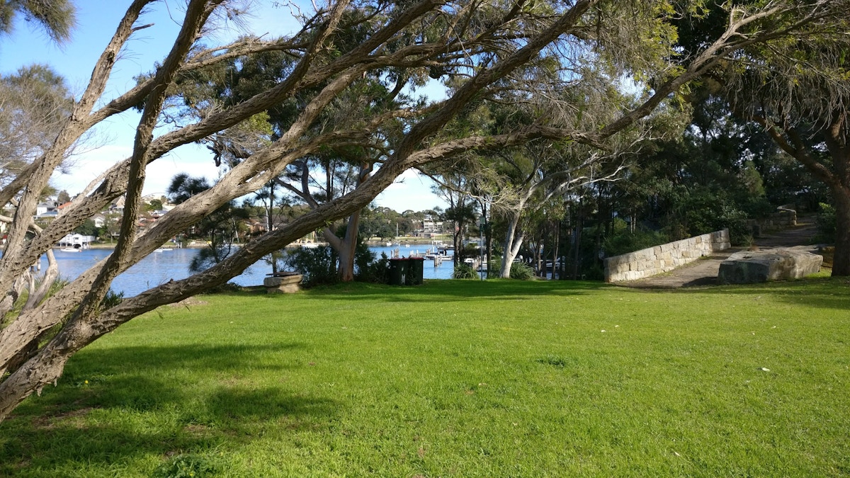 A grassy park with scattered trees overlooks a river with boats docked along the opposite shore under a partly cloudy sky.
