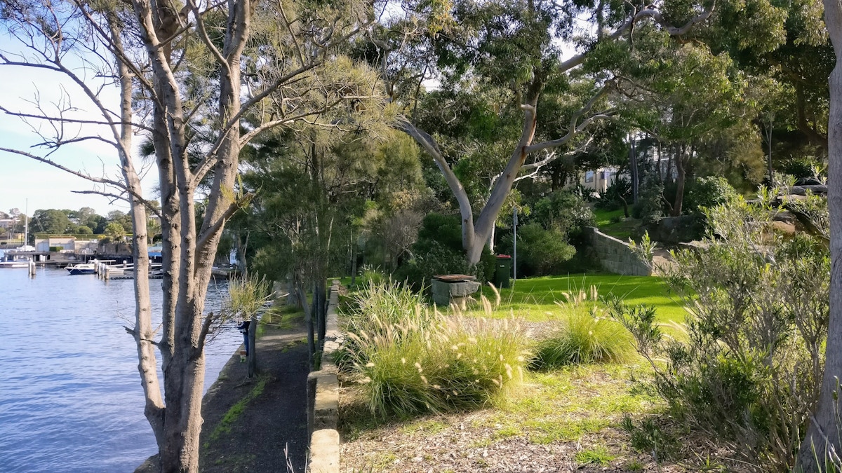 Trees and greenery line a grassy garden by the water, with a stone path and docked boats visible in the background under daylight.