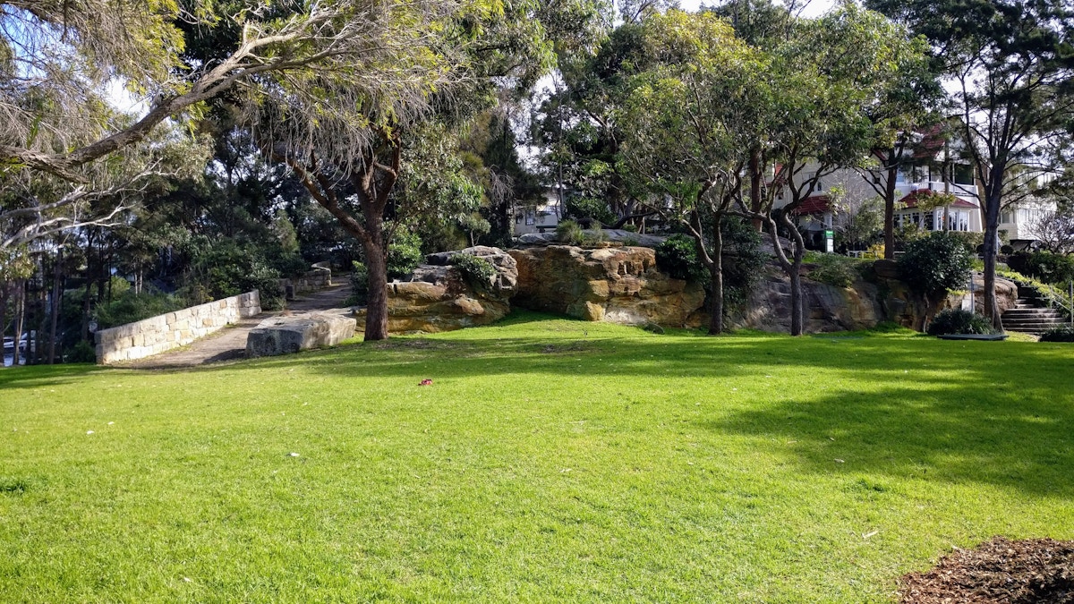 A grassy park area with scattered trees, large rocks, and a stone wall. Residential buildings are partially visible in the background.