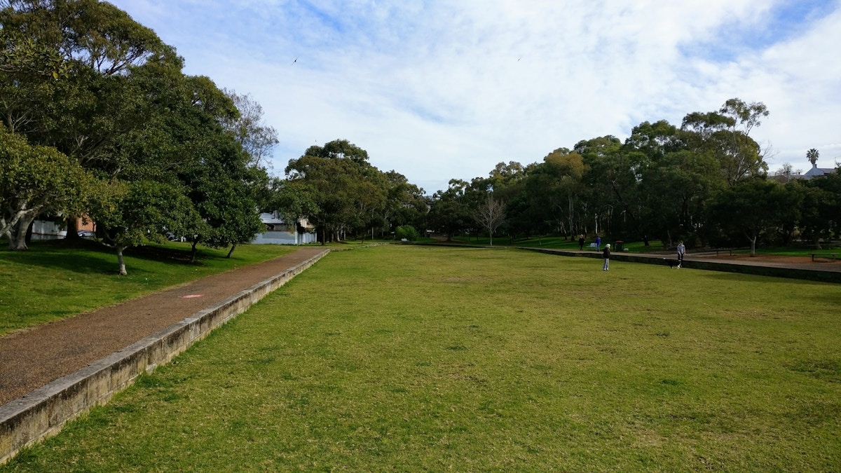 A large grassy park area with scattered trees, a paved walkway on the left, and a few people walking in the distance under a partly cloudy sky.