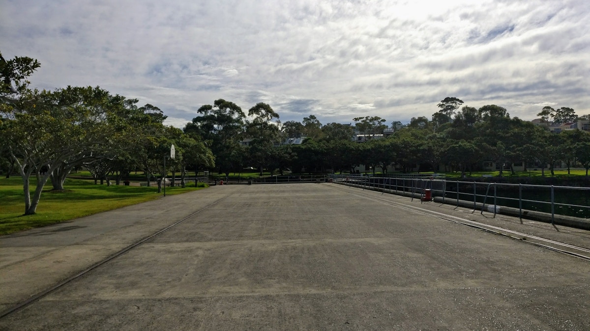 Wide paved path beside a river, bordered by green grass and trees, under a cloudy sky. Railings run along the river’s edge on the right.