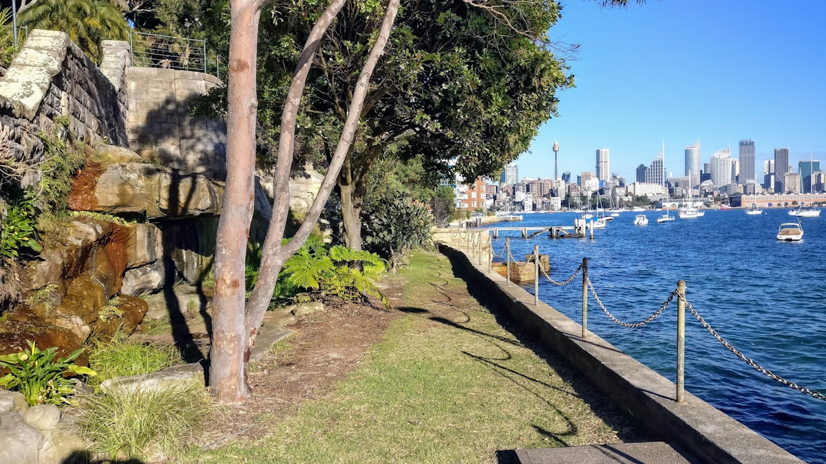 A waterfront path with trees and grass beside a stone wall overlooks a harbor with boats and a city skyline in the distance under a clear blue sky.