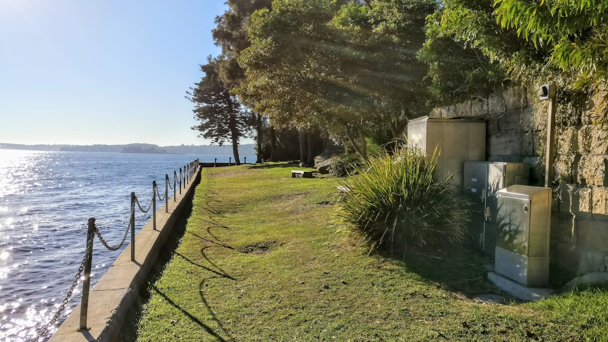 A grassy path runs beside a stone wall along a waterfront, with trees, utility boxes, and sunlight streaming through the branches.