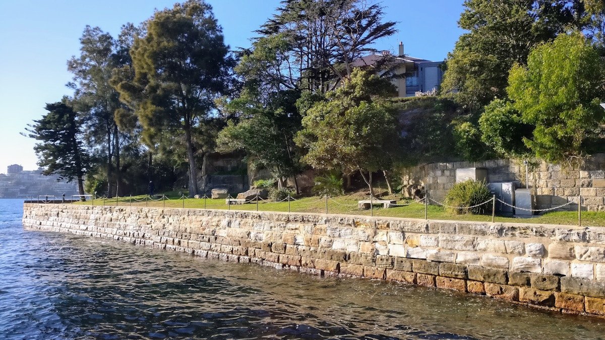 Stone seawall alongside clear water, with green trees and a house partially visible behind the foliage under a clear blue sky.