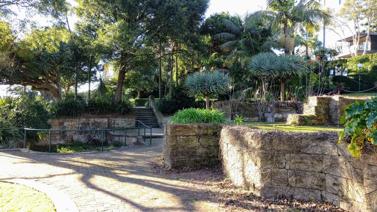 A stone path curves through a park with stone walls, stairs, lush greenery, tall trees, and sunlight filtering through the leaves.