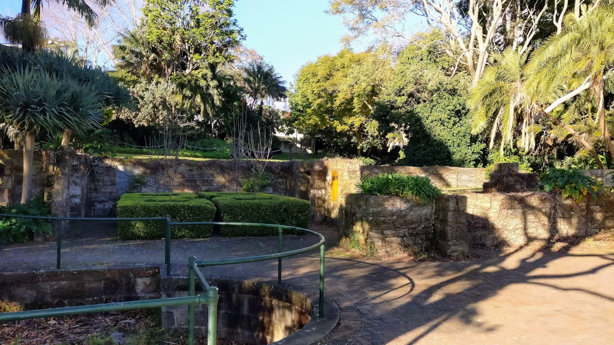 Sunlit garden with stone walls, trimmed hedges, and green metal railings, surrounded by various trees and shrubs.