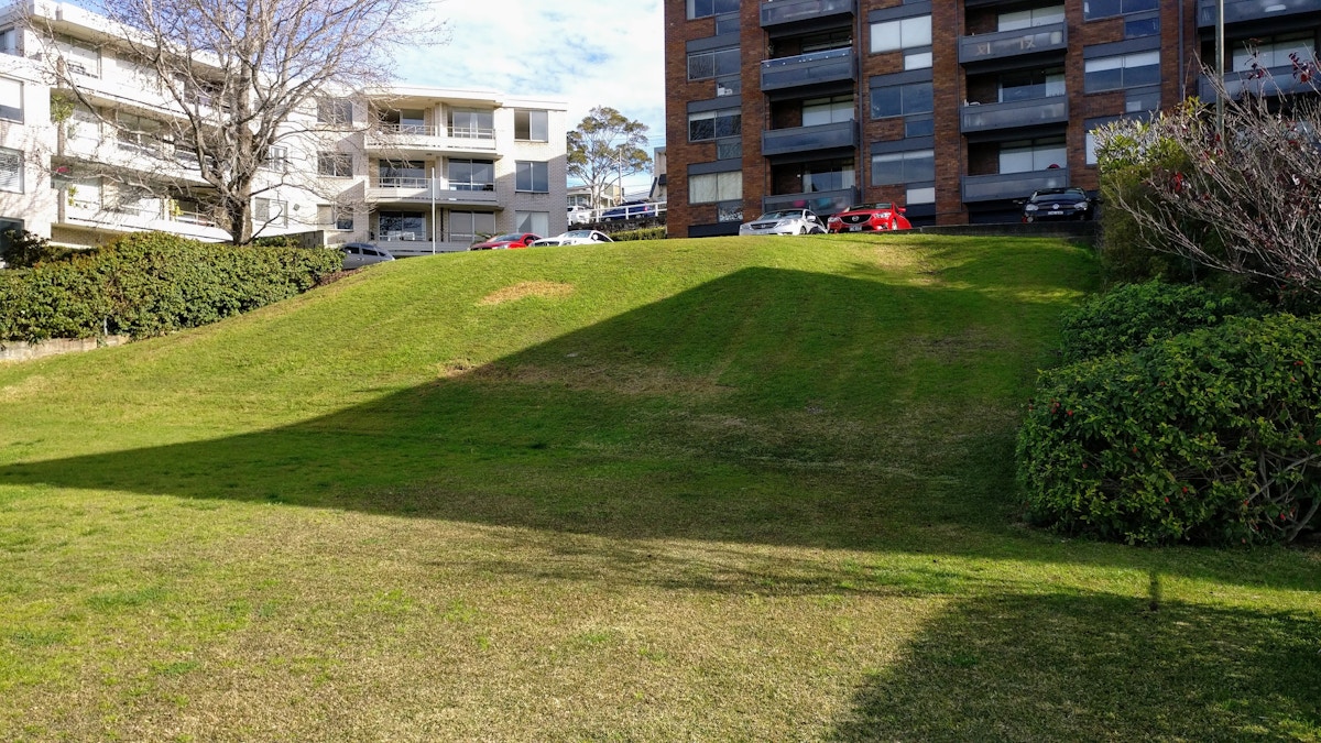 A grassy slope leads up to parked cars and apartment buildings under a partly cloudy sky. Bushes and trees border the lawn area.