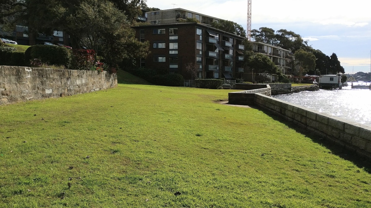 A grassy area slopes down toward a stone wall by the water, with apartment buildings and trees in the background.