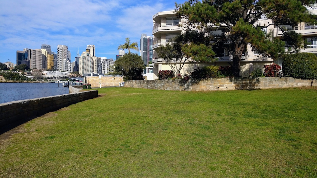 A grassy lawn leads to a waterfront with a modern apartment building on the right and city skyscrapers in the background under a blue sky.