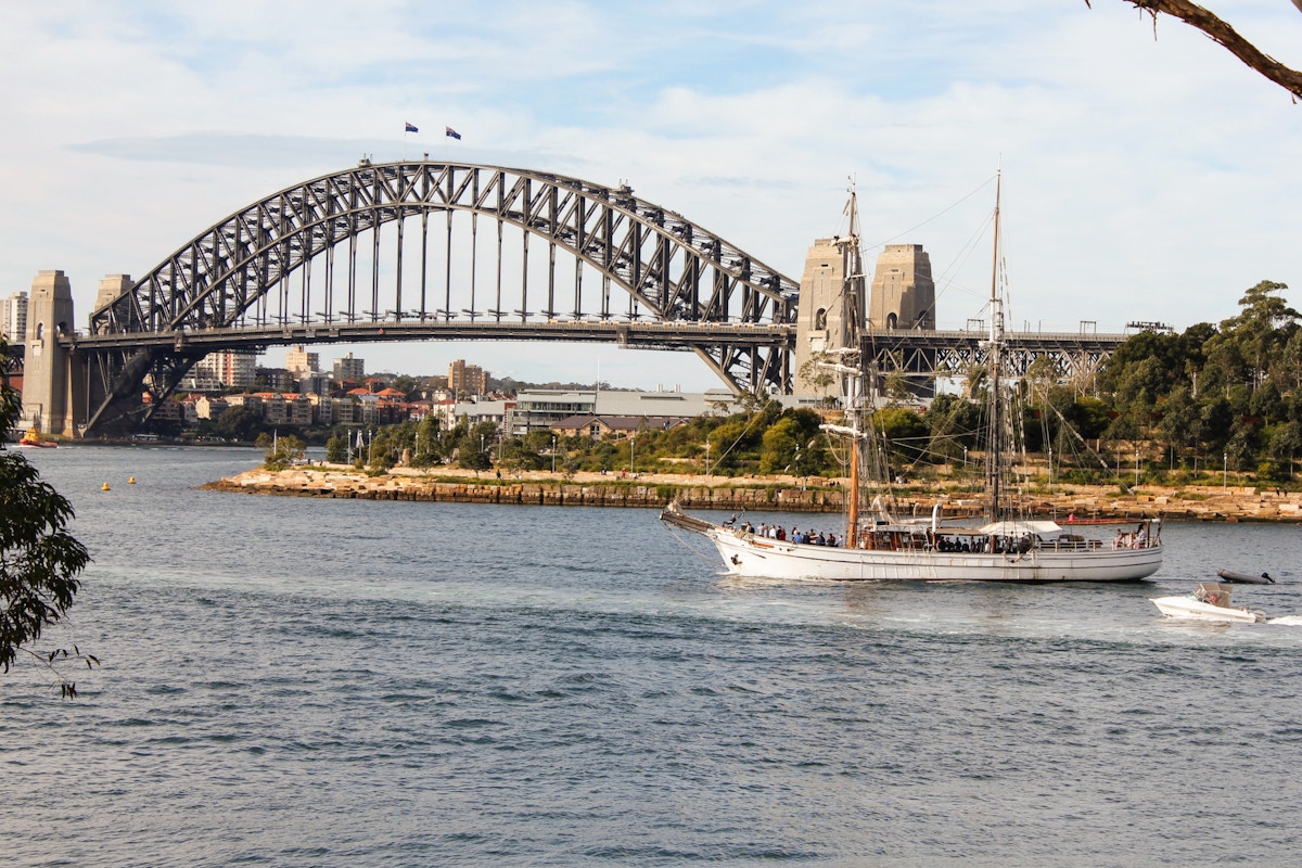 A white sailboat travels on the water near the Sydney Harbour Bridge, with city buildings and trees visible in the background under a partly cloudy sky.
