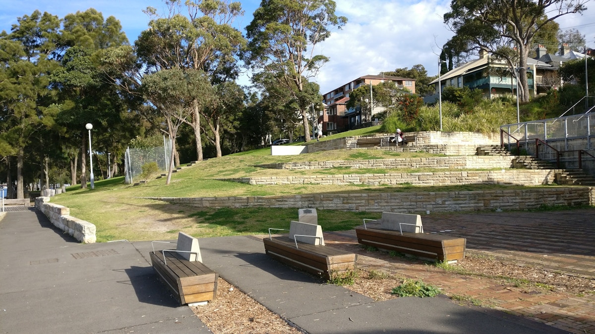 A small terraced park with wooden benches, stone retaining walls, trees, paths, and houses along the upper edge. Two people sit near the center on the stone seating steps.