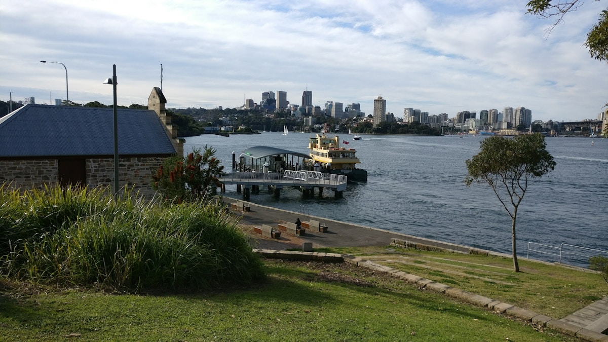 Ferry docked at a pier with grassy park in the foreground and city skyline in the background under a partly cloudy sky.