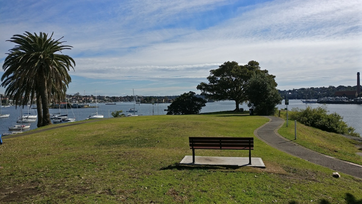 A wooden bench faces a harbor with sailboats, surrounded by grass, trees, a palm, and a paved path under a partly cloudy sky.