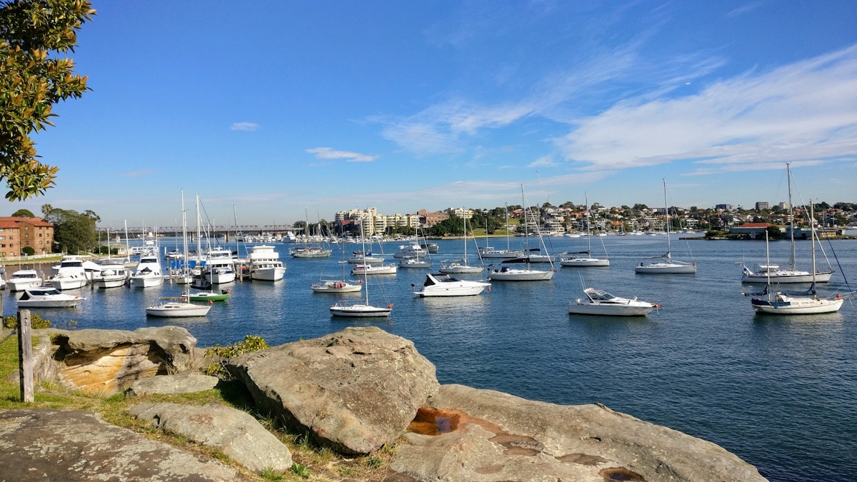 Numerous boats and yachts are anchored in a harbor with city buildings in the background and large rocks in the foreground under a blue sky.