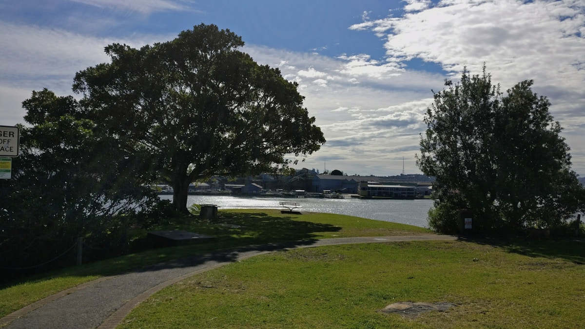A paved path curves past trees toward a waterfront, with boats and buildings visible across the water under a partly cloudy sky.