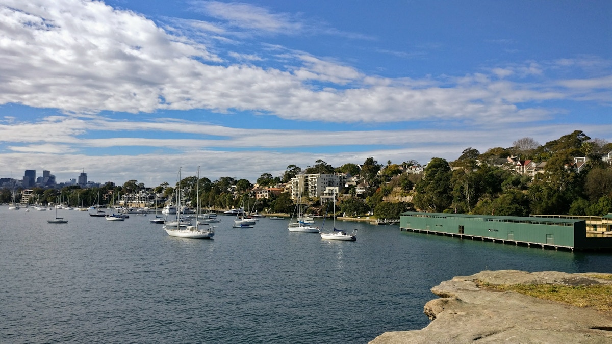 Several sailboats are anchored in a calm bay near residential buildings and a green boathouse, with trees and cloudy blue sky in the background.