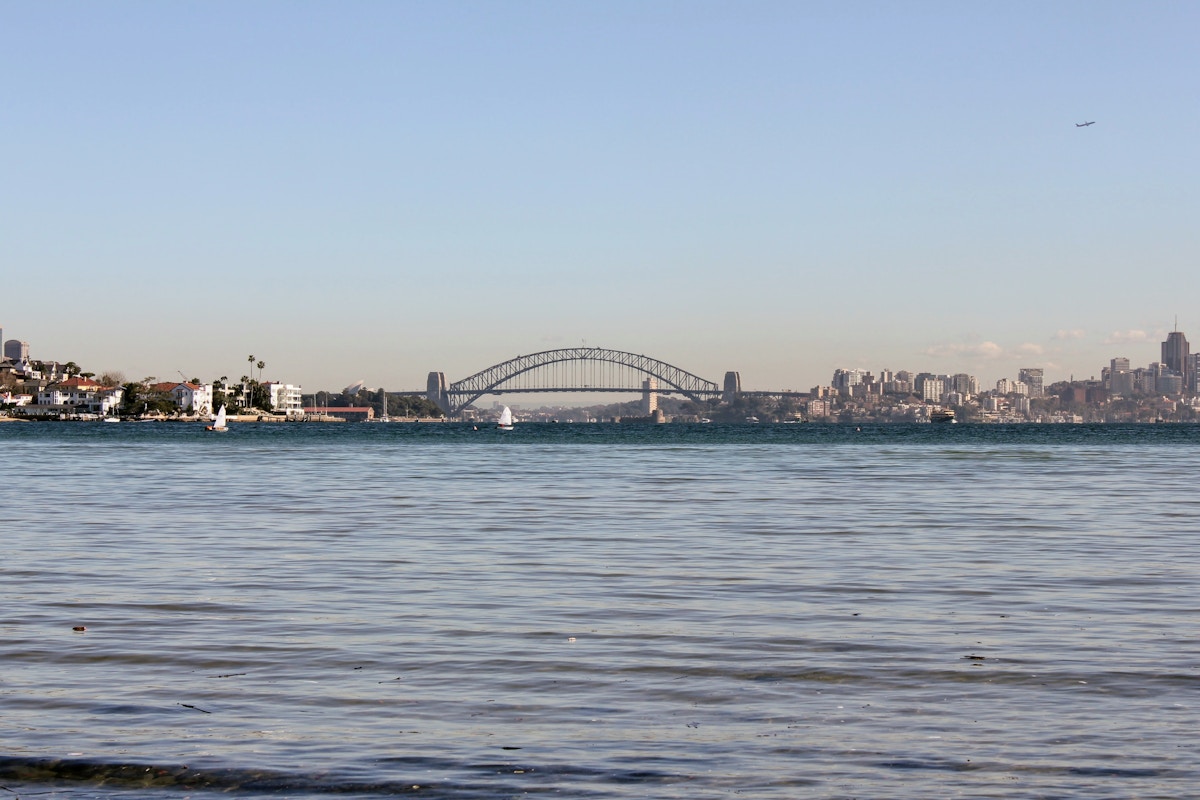 Sydney Harbour with the Sydney Harbour Bridge in the distance, city skyline on the right, and calm water in the foreground under a clear sky.