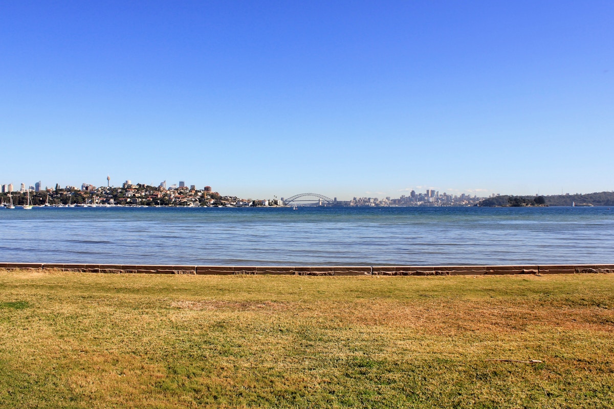 A grassy area leads to water with a city skyline and a large bridge visible in the distance under a clear blue sky.